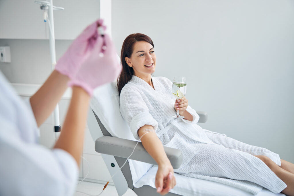 Woman smiling and holding a glass of water while she receives IV therapy