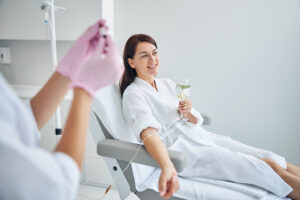 Woman smiling and holding a glass of water while she receives IV therapy